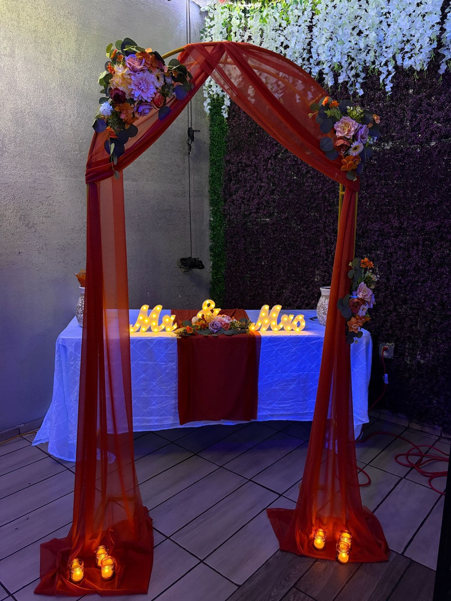 Sweetheart table under a terracotta-draped arch with florals and a lit Mr & Mrs sign