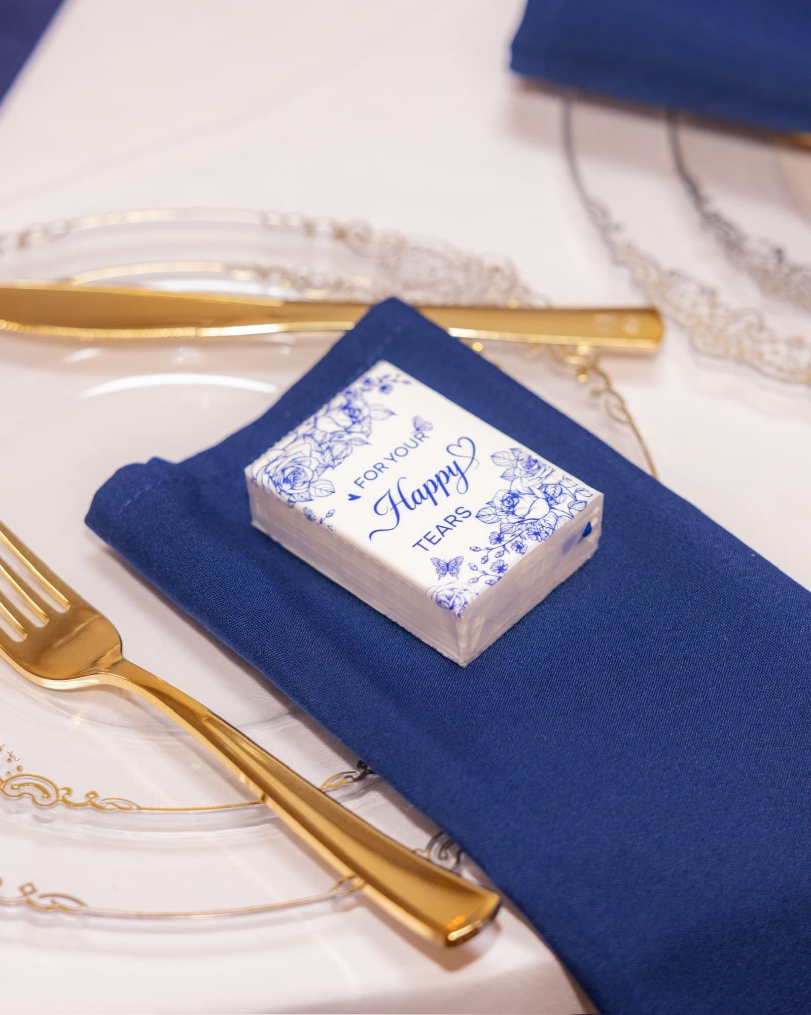 Place setting with a navy napkin, gold flatware, and 'Happy Tears' favor