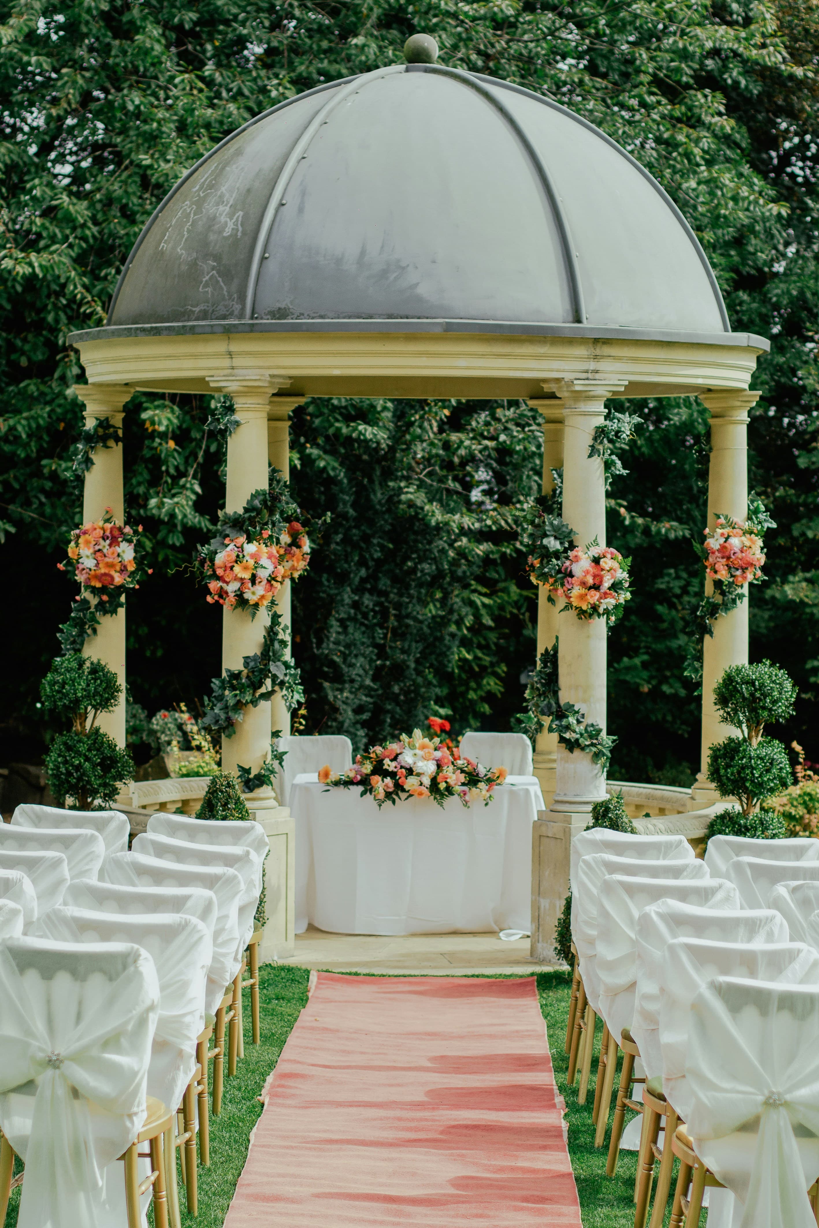 Una elegante ceremonia de boda al aire libre con una glorieta cubierta de flores y un pasillo en tono rosa